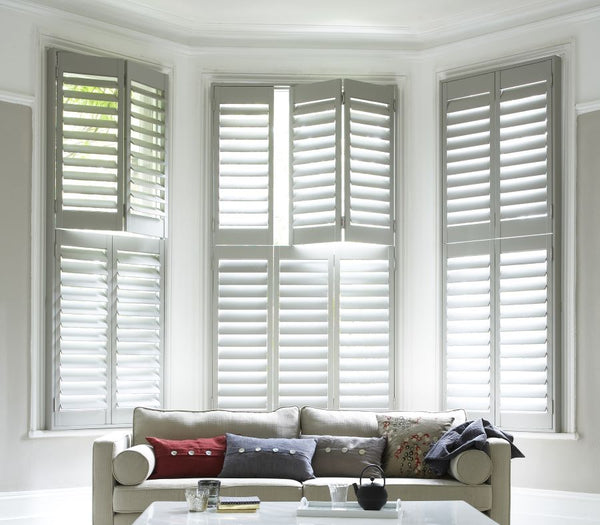 large bay window in a room dominated by white colours, with a white sofa and white large tier on tier wooden shutters